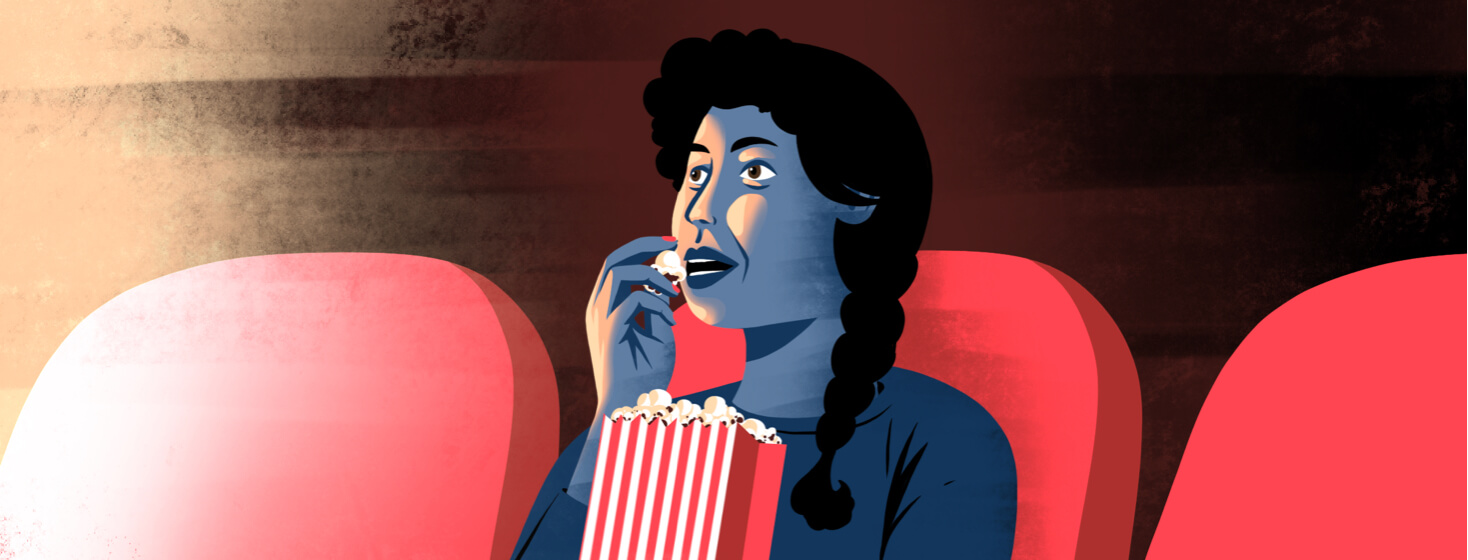 A woman is eating popcorn in a movie theater while a bright light from the screen shines on her face.